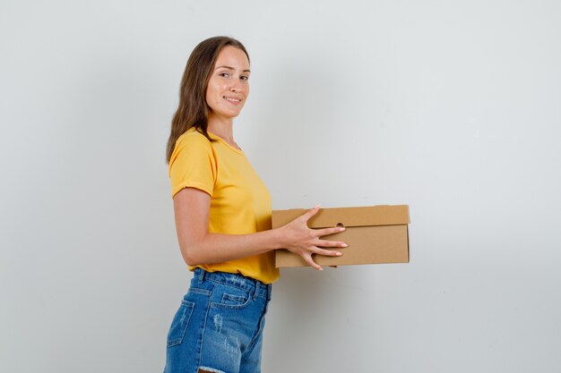 Young female in t-shirt, shorts holding cardboard box and smiling .