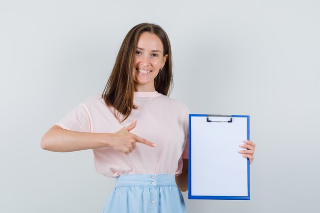 Young female in t-shirt, skirt pointing at clipboard and looking optimistic , front view.