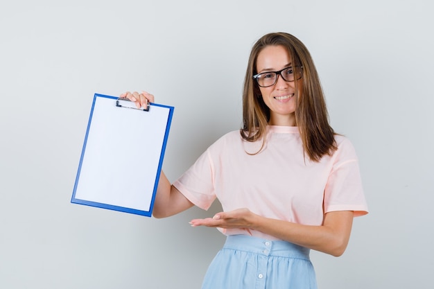 Young female in t-shirt, skirt showing clipboard and looking cheerful , front view.