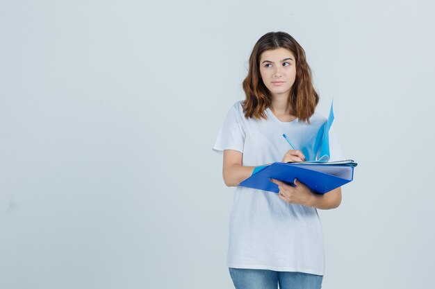 Young female taking notes on folder in white t-shirt, jeans and looking hesitant , front view.