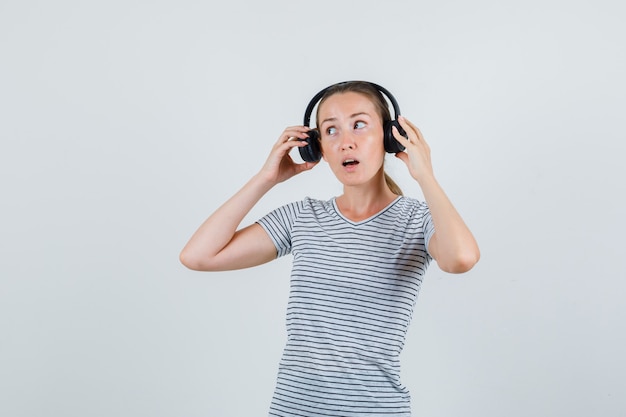 Young female taking off headphones in striped t-shirt and looking curious , front view.