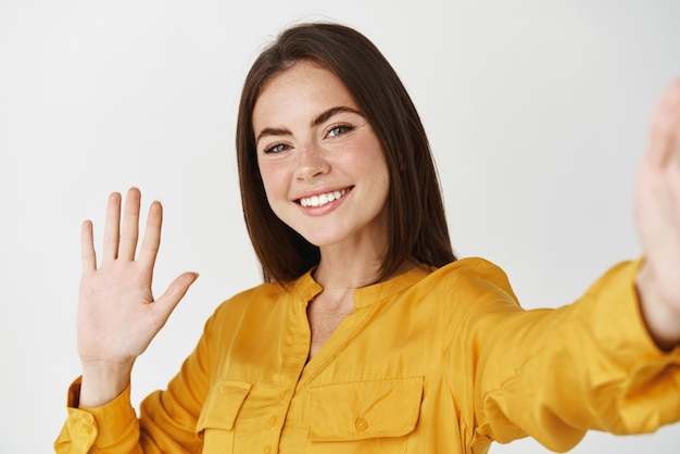 Young female taking selfie recording herself on smartphone Woman waving hand at camera during video call standing over white background