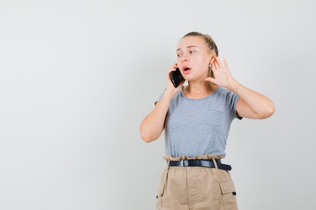 Young female talking on mobile phone, holding hand behind ear in t-shirt, pants , front view.