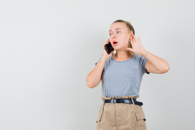 Tシャツ、ズボン、正面図で耳の後ろに手をつないで、携帯電話で話している若い女性。