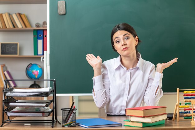 young female teacher sitting at table with school tools in classroom