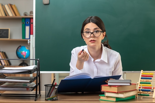 교실에서 학교 도구와 함께 테이블에 앉아 폴더를 들고 안경을 쓴 젊은 여교사