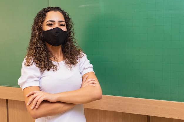 팔짱을 낀 젊은 여교사는 새로운 표준의 수술용 마스크를 쓰고 칠판을 배경으로 교실을 건넜다. 전염병 후 학교로 돌아가는 개념 프리미엄 사진