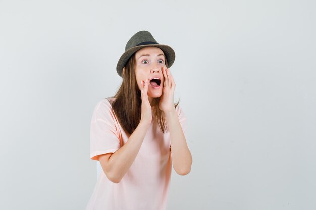 Young female telling secret in pink t-shirt, hat and looking excited. front view.