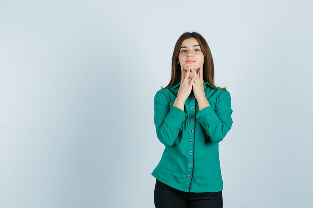 Young female touching her chin with fingers in green shirt, pants and looking cute , front view.