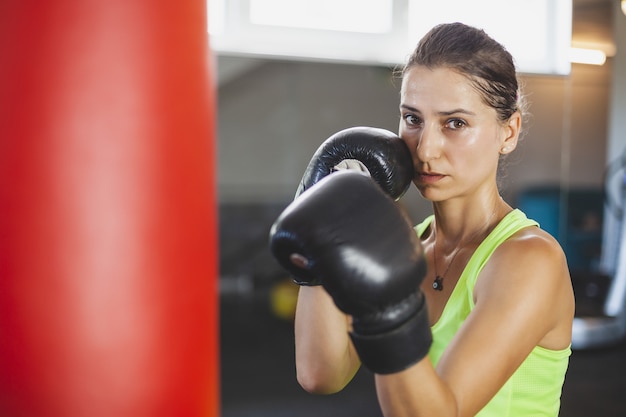 若い女性はジムでトレーニングし、サンドバッグの前でボクシンググローブでボクシングの練習をします