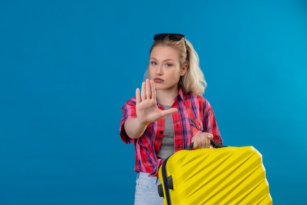 Young female traveler wearing red shirt and glasses on head holding suitcase showing stop gesture on isolated blue wall