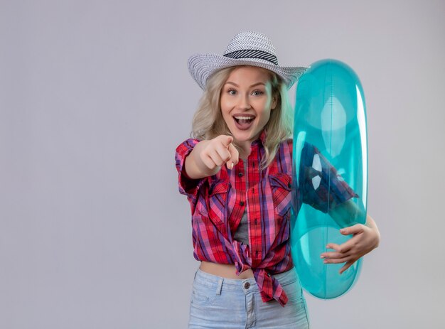 Young female traveler wearing red shirt in hat holding inflatable ring showing you gesture on isolated white wall