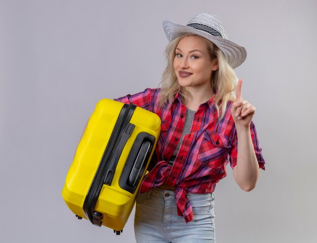 Young female traveler wearing red shirt in hat holding suitcase points to up on isolated white wall