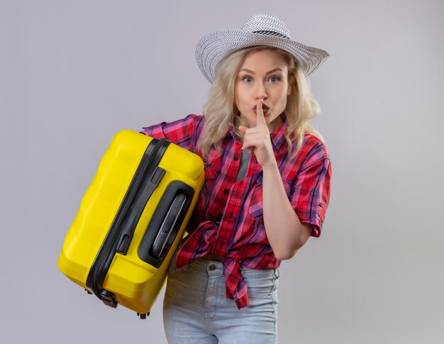 Young female traveler wearing red shirt in hat holding suitcase showing silence gesture on isolated white wall