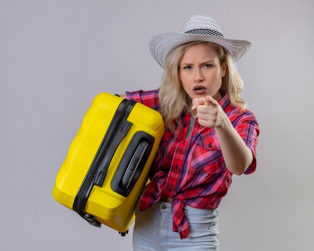 Young female traveler wearing red shirt in hat holding suitcase showing you gesture on isolated white wall