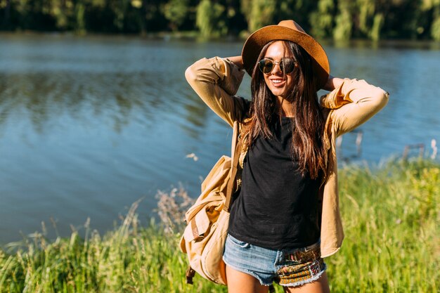 Young female traveler with backpack, glasses and hat standing in front of lake