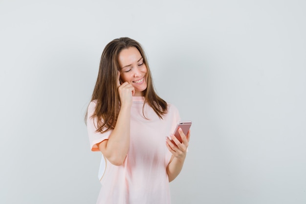 Giovane femmina utilizzando il telefono cellulare in maglietta rosa e guardando allegra, vista frontale.