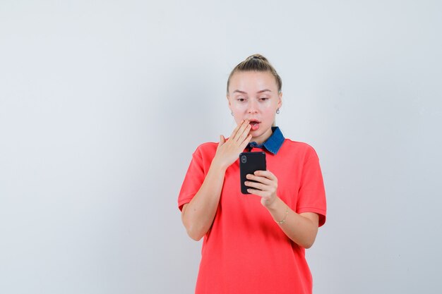 Young female using mobile phone in t-shirt and looking surprised , front view.