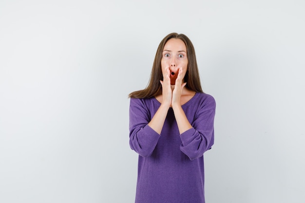 Young female in violet shirt holding hands near open mouth and looking scared , front view.