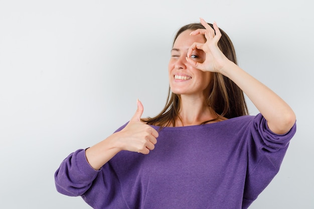 Foto gratuita giovane femmina in camicia viola che mostra gesto giusto con il pollice in su e che sembra allegra, vista frontale.