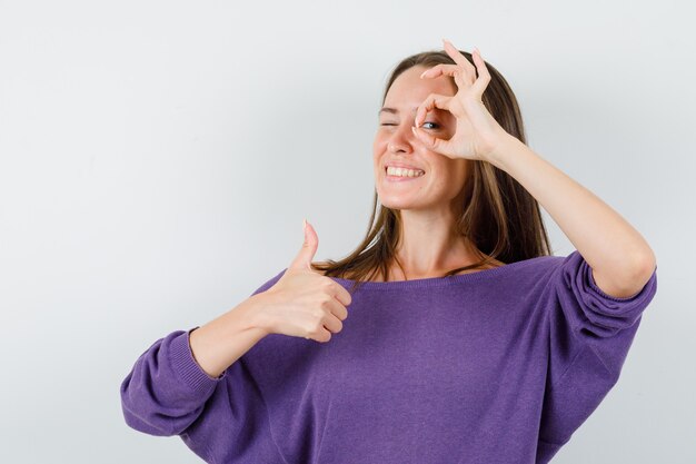 Young female in violet shirt showing ok gesture with thumb up and looking cheerful , front view.