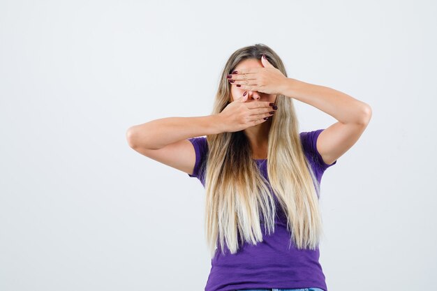 Молодая женщина в фиолетовой футболке, взявшись за руки на глазах и во рту и глядя скрытно, вид спереди.