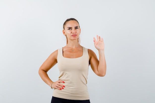 Young female waving hand for greeting while keeping hand on hip in beige tank top and looking confident