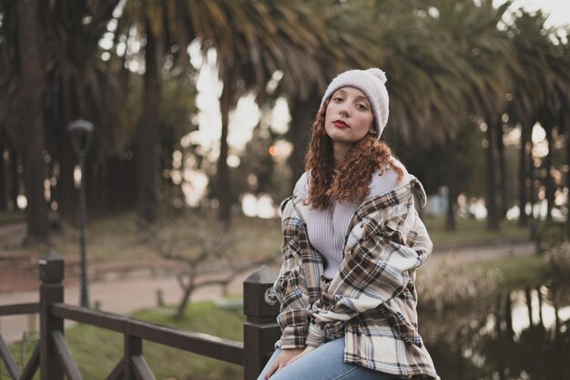 Young female wearing a plaid jacket and a white hat sitting on the wooden fence in the park