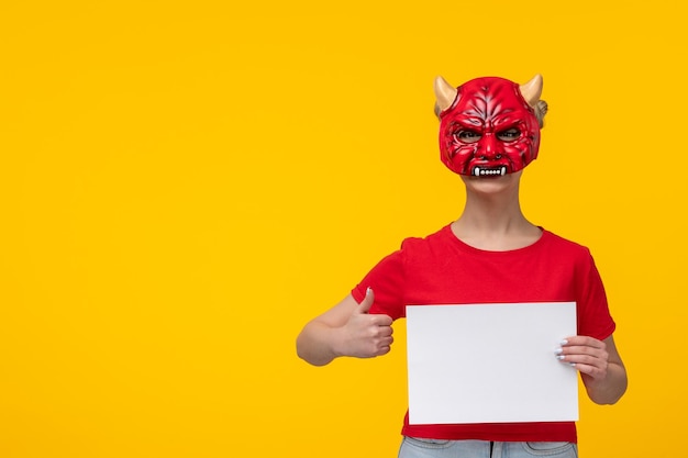 Young female wearing scary devil mask holding piece of paper yellow background ugly