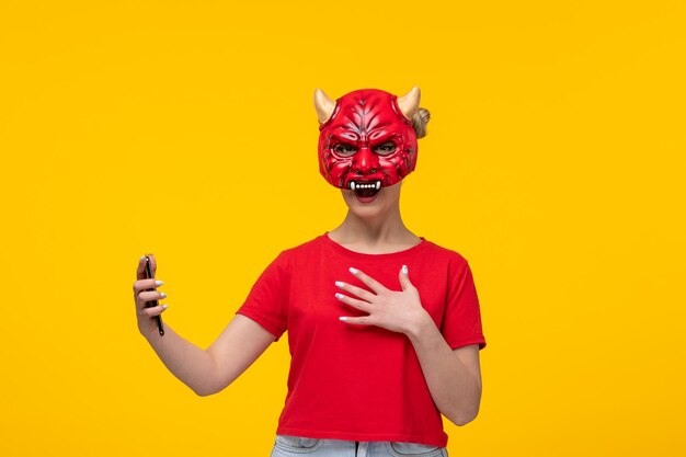 Молодая женщина в страшной маске дьявола с помощью своего телефона на желтом фоне уродлива