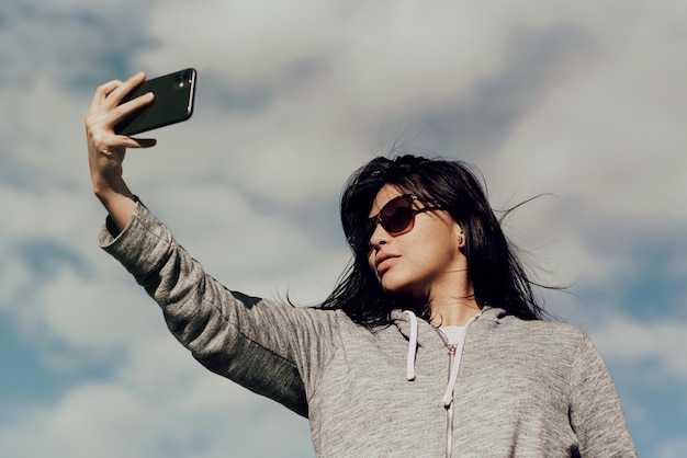 曇った青い空の下で彼女の携帯電話で写真を撮るサングラスをかけている若い女性