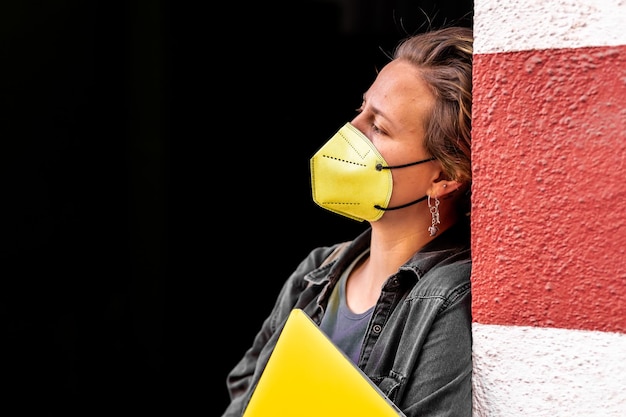 Free photo young female wearing a yellow face mask and holding a laptop leaning on a wall