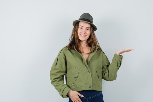 Young female welcoming or showing something in jacket, pants, hat and looking merry , front view.