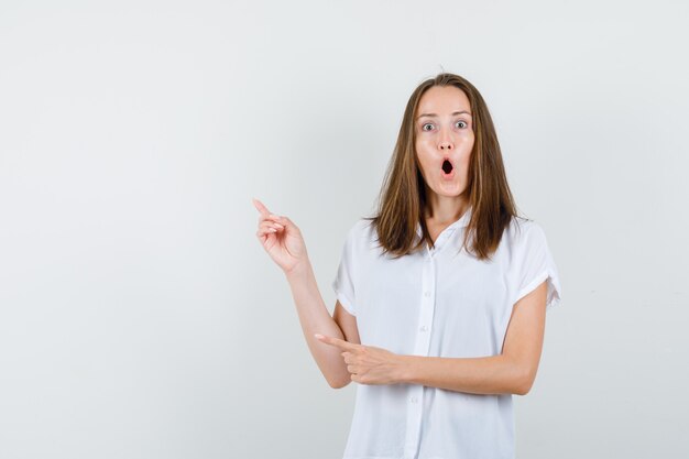 Young female in white blouse pointing aside and looking astonished