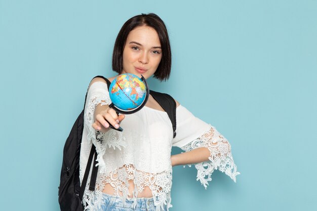 молодая женщина в белой рубашке и черной сумке позирует и держит глобус на синем
