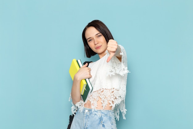 young female in white shirt blue jeans and black bag holding copybooks on blue