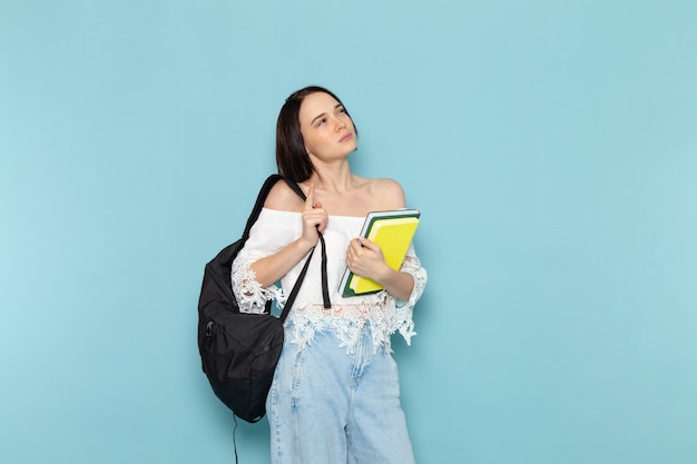 young female in white shirt blue jeans and black bag holding copybooks on blue