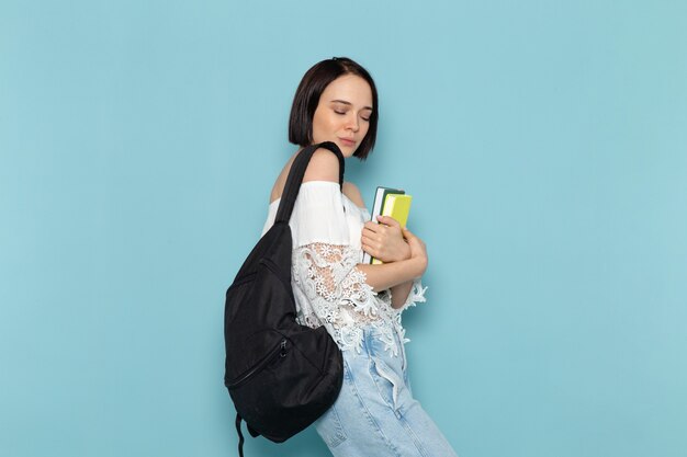 young female in white shirt blue jeans and black bag holding copybooks on blue