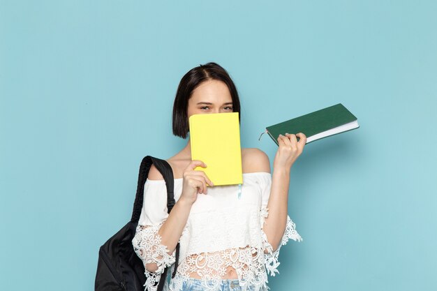 young female in white shirt blue jeans and black bag holding copybooks on blue