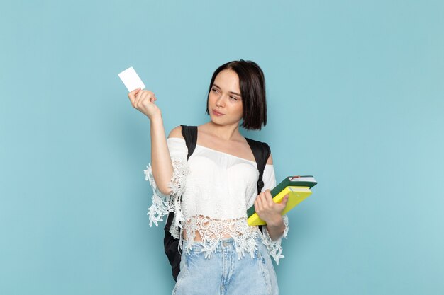 young female in white shirt blue jeans and black bag holding copybooks and card on blue