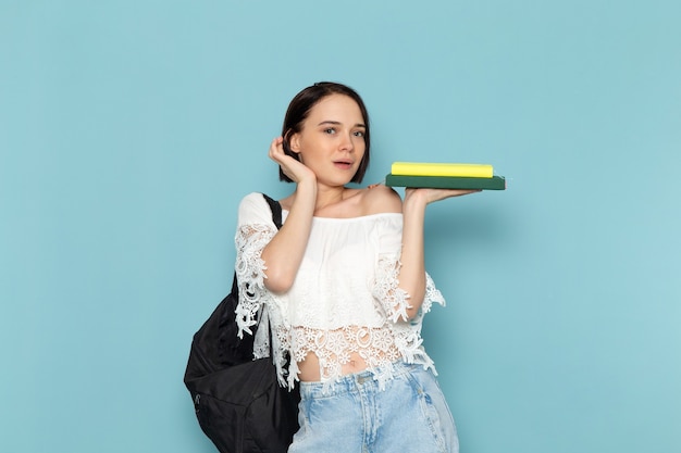 young female in white shirt blue jeans and black bag holding copybooks posing on blue