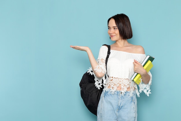 young female in white shirt blue jeans and black bag holding copybooks smiling on blue