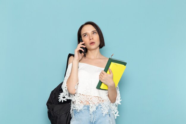 young female in white shirt blue jeans and black bag holding copybooks talking on phone on blue