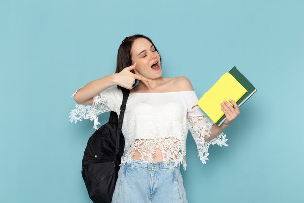 young female in white shirt blue jeans and black bag holding copybooks with smile on blue