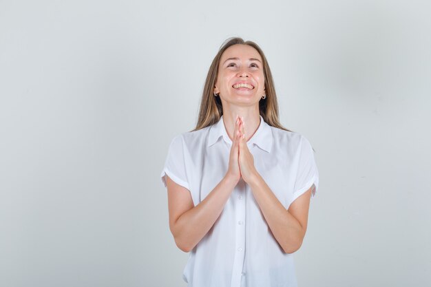 Young female in white shirt keeping hands in praying gesture and looking happy