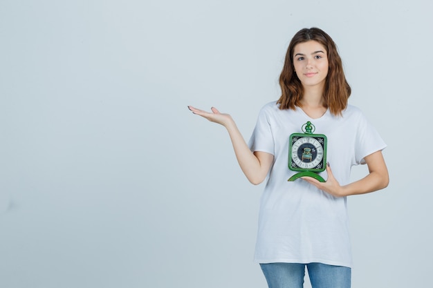 Free photo young female in white t-shirt, jeans holding clock, showing welcoming gesture and looking cheery , front view.