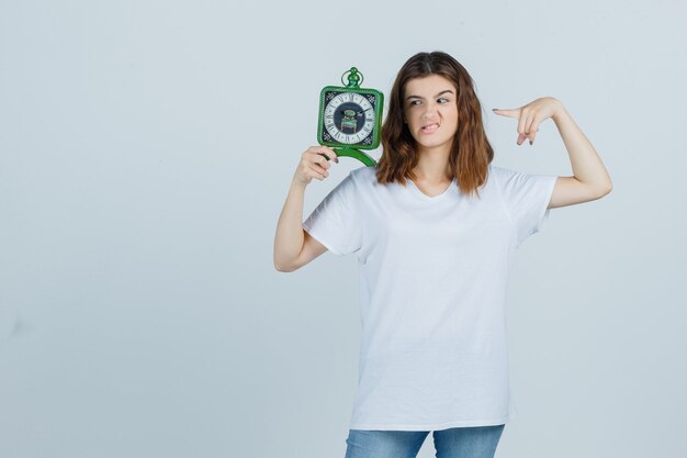 Молодая женщина в белой футболке, джинсы, указывающие на часы, хмурое лицо и обеспокоенный вид, вид спереди.
