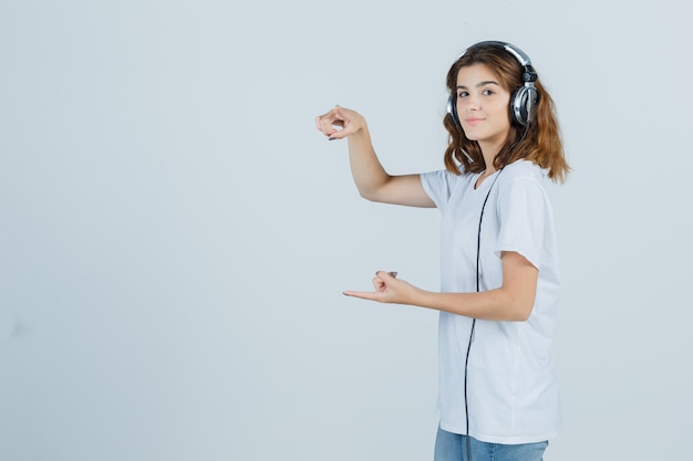 Young female in white t-shirt pointing fingers to the front and left side and looking merry , front view.