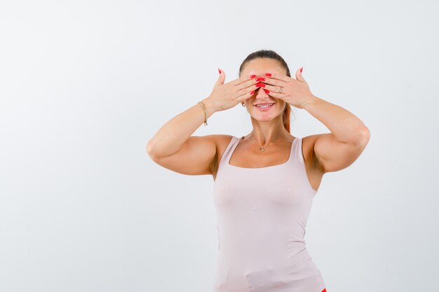 Young female in white tank top holding hands on eyes and looking cute , front view.