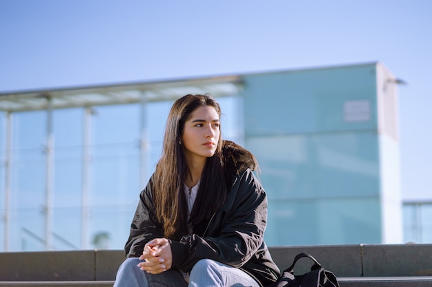 Young female with a black jacket sitting on concrete stairs with clenched hands looking aside